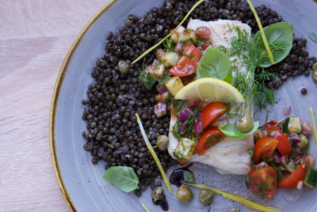 Close-up of a delicious cod fish dish with lentils, salsa, and fresh herbs on a plate.