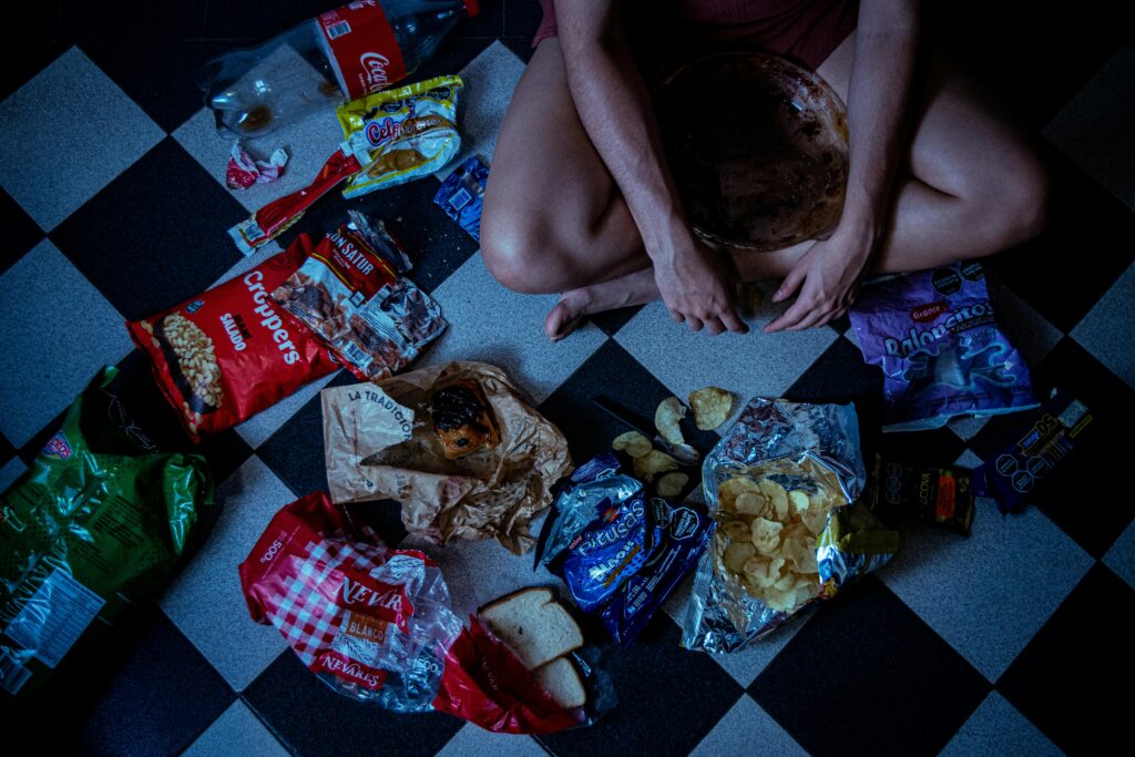 Man sitting surrounded by assorted snacks and packaging on a checkered floor, depicting a binge-eating scene.